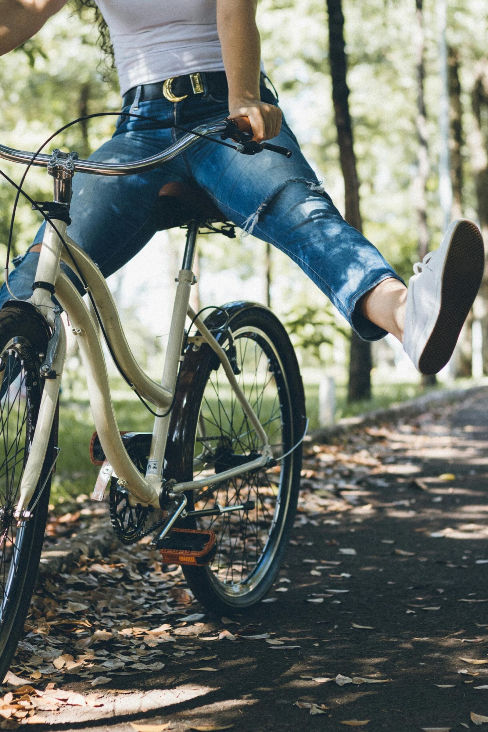 Person in jeans and white sneakers sitting on a light-colored bicycle on a leaf-covered forest path.