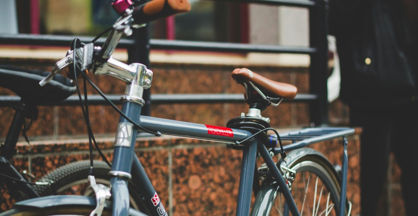 Close-up of a dark gray city bike with brown saddle, parked near a stone building with a person in the background.
